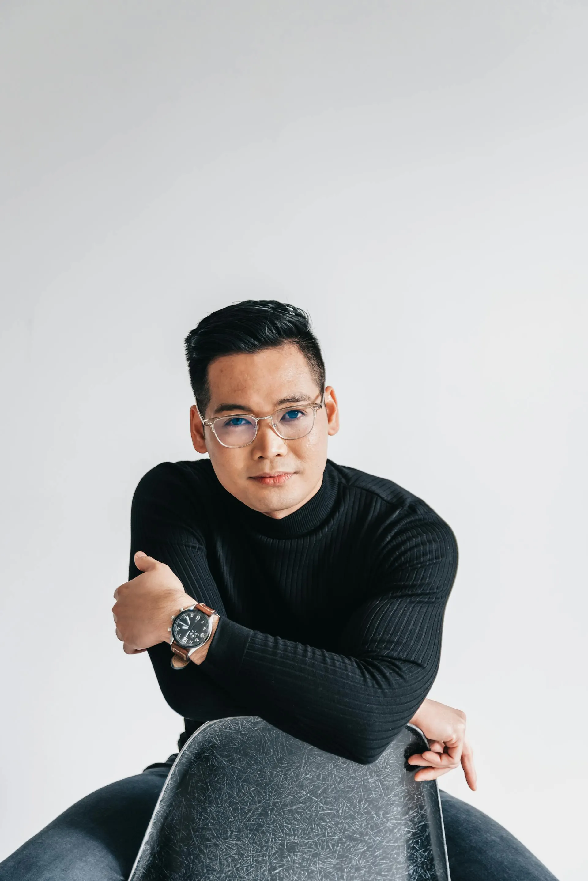 Young man wearing glasses and a black turtleneck sweater sitting on a chair against a plain white background.
